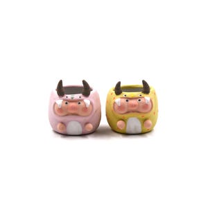 May include: Two ceramic planters in the shape of cartoon monsters. One is pink with brown horns, and the other is yellow with brown horns. Both have a pig-like snout and small arms and legs. These planters are ideal for small succulents or cacti.