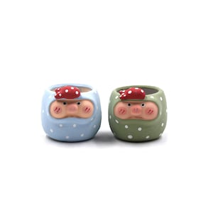 May include: Two ceramic planters, each shaped like a pig with a red polka-dot head covering. One planter is light blue with white polka dots, the other is green with white polka dots. The planters have a round shape and are suitable for small plants.