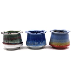 May include: Three ceramic tea strainers with flared, scalloped rims. The left strainer has a green, red, and white glaze. The center strainer features a deep blue glaze with white drips. The right strainer displays a blue, yellow, and red gradient.