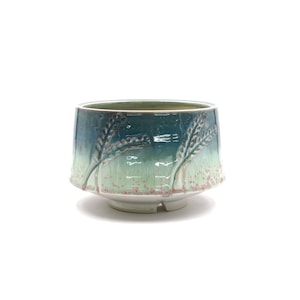 Può includere: Una ciotola in ceramica con una glassa verde e blu. La ciotola presenta un design in rilievo di spighe di grano sui lati. La ciotola ha una base bianca.