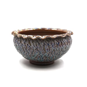 May include: A decorative ceramic bowl with a textured, mottled surface in shades of blue, brown, and green. The rim has a scalloped edge with a brown and gold trim. The bowl sits on a small, round base.