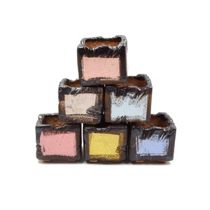 Puede incluir: Un conjunto de seis pequeñas macetas cuadradas de terracota con esmaltes de diferentes colores. Las macetas están apiladas en forma de pirámide, con la maceta más grande en la parte inferior y la más pequeña en la parte superior. Los esmaltes son rosa, azul claro, amarillo y azul oscuro.