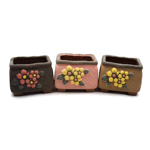 May include: Three small square ceramic flower pots with different colored glazes. The pots have a brown rim and are decorated with painted flowers. The pots are arranged in a row from left to right: brown, pink, and brown.