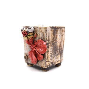 May include: A ceramic planter with a whimsical design featuring a red and yellow fish with large eyes. The planter has a textured, aged look and is a unique addition to any home.