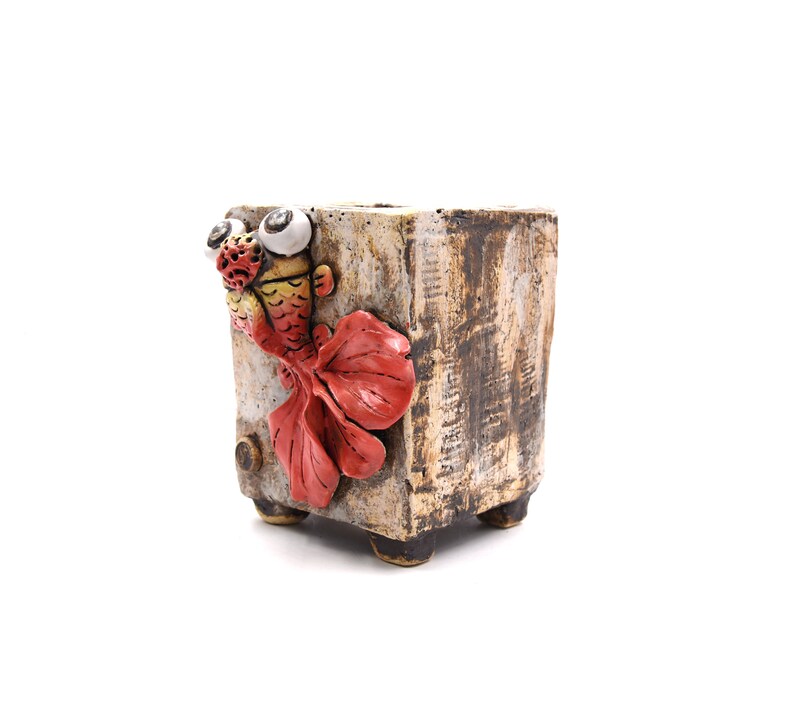 May include: A ceramic planter with a whimsical design featuring a red and yellow fish with large eyes. The planter has a textured, aged look and is a unique addition to any home.