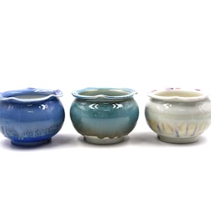 May include: Three small ceramic bowls with scalloped rims and rounded bodies. The first bowl is deep blue with a glossy finish, the second is a teal green with a subtle crackle glaze, and the third is a pale cream with soft purple accents.