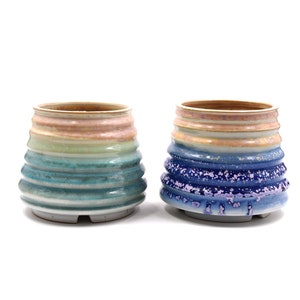 以下が含まれることがあります： テクスチャ加工されたリブ付きのデザインのセラミックプランター2つ。左側のプランターは青、緑、ピンクの配色。右側のプランターは青、ピンク、白の配色です。