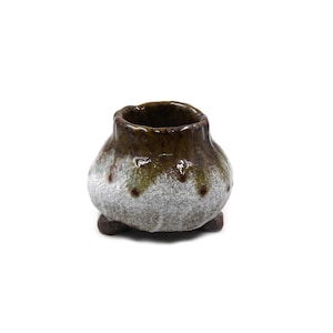 May include: A small, round ceramic pot with three short feet. The upper portion features a glossy, dark brown glaze that drips down over a textured, matte white and light gray base. The rim is uneven and circular, creating a wide opening.