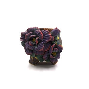 May include: A decorative ceramic planter with a floral design. The planter features three-dimensional purple flowers with red edges and yellow centers. The leaves are green. The planter is brown and has three small feet.