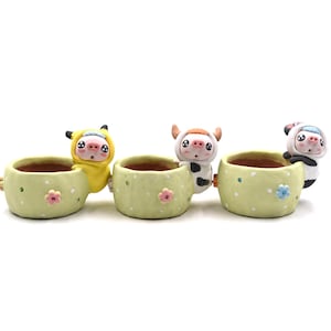 May include: A set of three light green ceramic planters, each adorned with a cartoon animal figure. The planters are decorated with small flowers and polka dots. The animal figures include a yellow pig, a cow, and a panda.