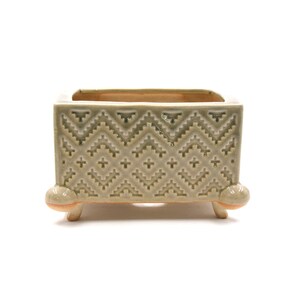 May include: A square ceramic planter in shades of green and beige, with a geometric pattern. The planter has a rectangular opening and is supported by three small, rounded feet. The design features a zig-zag pattern.