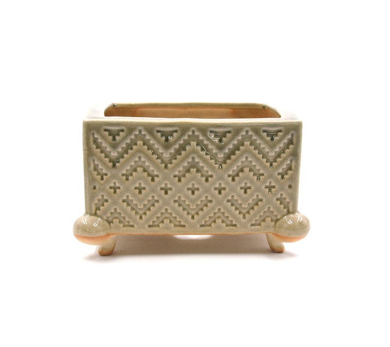 May include: A square ceramic planter in shades of green and beige, with a geometric pattern. The planter has a rectangular opening and is supported by three small, rounded feet. The design features a zig-zag pattern.