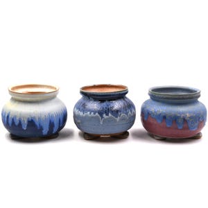 May include: Three small, round ceramic pots with a glazed finish. Each pot has a different color scheme, including blue, white, and red hues. The pots have a small, rounded base and a contrasting interior color.