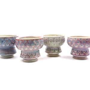 May include: Four ceramic bowls with a pedestal base, each with a unique color scheme. The bowls feature a repeating pattern of small, stylized flowers. Colors include blue, red, green, and purple, with a glossy finish.