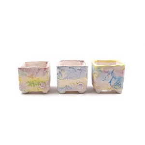May include: Three small square ceramic planters with a colourful, marbled glaze. Each planter has a small, round, white button on the front.