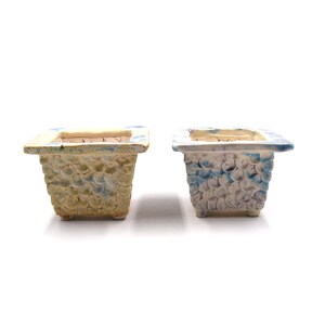 Puede incluir: Dos macetas cuadradas de cerámica con patrones texturizados en forma de escamas. Una es principalmente beige con detalles azules y verdes, la otra es blanca con detalles azules. Ambas tienen un borde cuadrado y se asientan sobre pequeños pies.