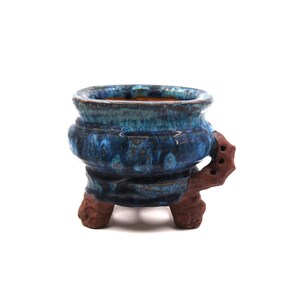 Puede incluir: Pequeña maceta de cerámica artesanal con acabado esmaltado turquesa y azul. La maceta tiene una forma única con una amplia abertura y está soportada por tres patas marrones texturizadas. Diseñada para plantas pequeñas.