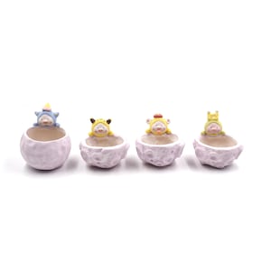 May include: Four small, pastel purple ceramic planters shaped like the moon, each with a different animal figurine peeking out of the top. The animals are a blue unicorn, a yellow Pikachu, a yellow bear, and a green frog.