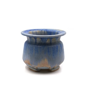 May include: A small, blue ceramic pot with a rounded body and a flared rim. The pot has a textured surface with streaks of lighter blue and tan. It sits on three small feet.