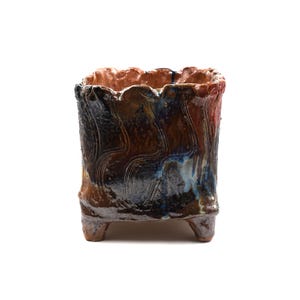 May include: A handcrafted ceramic planter with a square shape and three small feet. The exterior features a textured, swirling pattern in shades of brown, black, and blue, with a reddish-brown rim.