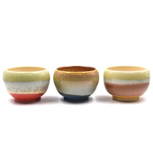 May include: Three ceramic bowls with a round shape and a colourful, layered design. The bowls feature a mix of colours, including yellow, white, red, brown, and blue. The bowls are likely for serving food or as decorative items.