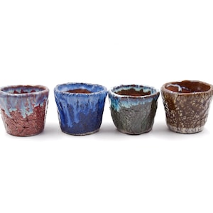 Op de afbeelding: Vier kleine keramische potten in verschillende kleuren en patronen. De potten zijn in een rij van links naar rechts gerangschikt. De pot links is rood met blauwe glazuur, de volgende is blauw met witte glazuur, de volgende is groen met bruine glazuur en de laatste is bruin met witte glazuur.