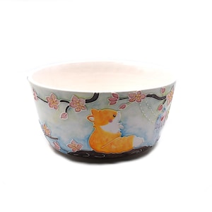 May include: A ceramic bowl with a light blue exterior featuring a cartoon fox and floral design. The bowl has a white interior and a dark brown base. Pink flowers and green leaves decorate the bowl's exterior.
