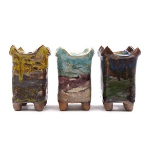 May include: Three small, square ceramic pots with a unique, hand-painted glaze. Each pot has a different color scheme, including yellow, brown, blue, green, and purple. The pots have four small feet and are perfect for displaying small plants or succulents.