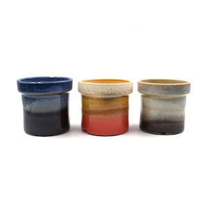 May include: Three ceramic planters with a cylindrical shape and a wide rim. The planters feature a gradient color scheme, with shades of blue, red, and brown. The planters are suitable for small plants or succulents.