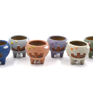 May include: Six small ceramic planters with a cartoon city skyline design. The planters are in various colors, including blue, purple, green, and orange. Each planter has three small legs and a cloud design.