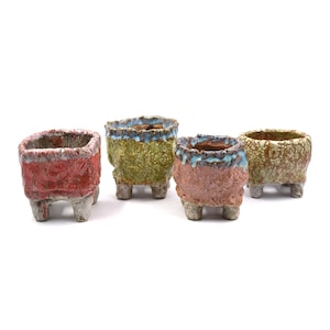 May include: Four small, textured ceramic planters in shades of red, green, brown, and tan. Each planter has a unique, rough surface and sits on three small, rounded feet. The planters have a rustic, handmade appearance.