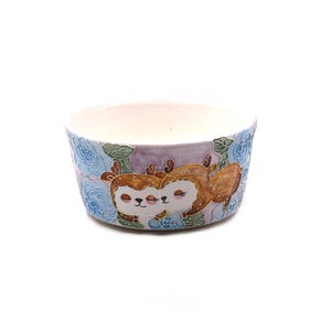 Puede incluir: Un cuenco de cerámica con interior lila claro y exterior pintado. El exterior presenta dos ciervos abrazándose, rodeados de diseños florales azules. Los ciervos son marrones con detalles blancos y ojos cerrados. El cuenco es redondo y tiene fondo blanco.