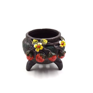 Peut inclure: Un petit pot en céramique noire avec trois pieds et un motif de fraises rouges et de fleurs jaunes. Le pot est parfait pour les petites plantes ou les succulentes.
