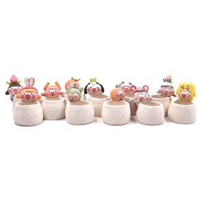 May include: A collection of ten small, speckled off-white ceramic planters, each featuring a unique miniature pig figurine perched on the rim. The pigs are decorated with various colorful headpieces, including fruits, vegetables, and animal ears.