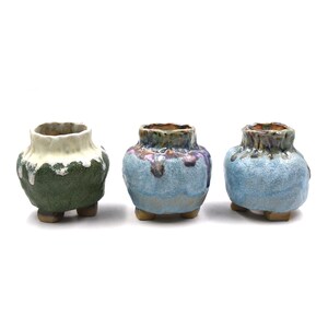 May include: Three small ceramic pots with textured surfaces and three short feet each. The left pot is dark green with a white drip glaze. The middle and right pots feature light blue and purple mottled glaze patterns with brown rim details.