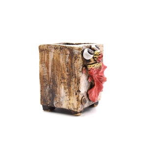 May include: A brown ceramic planter with a whimsical fish design. The fish has red fins, yellow scales, and white eyes.