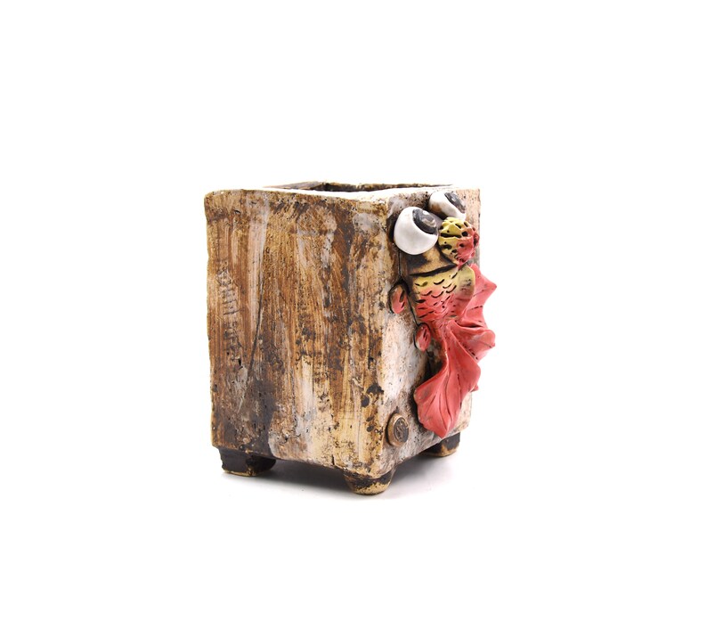 May include: A brown ceramic planter with a whimsical fish design. The fish has red fins, yellow scales, and white eyes.