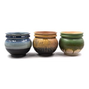 May include: Three ceramic pots with rounded bodies and flared rims. The first pot features a blue and black gradient glaze. The second pot has a yellow and brown drip glaze. The third pot displays a green and cream drip glaze finish.