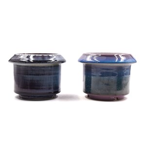 Pode incluir: Dois vasos de cerâmica com um acabamento único de esmalte. O vaso à esquerda tem um esmalte azul escuro e roxo com uma borda branca. O vaso à direita tem um esmalte azul e roxo com uma borda rosa.