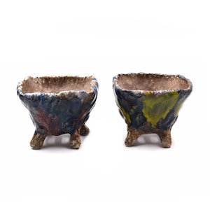 May include: Two small, square ceramic pots with a textured, uneven rim. The pots are supported by three small feet. One pot has blue, red, and brown hues, while the other has blue and yellow tones. The interior of both pots is a light brown color.