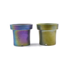 May include: Two small, cylindrical planters with a flared rim. One planter has a metallic, iridescent finish with hues of blue, purple, and gold. The other planter is a solid olive green color. Both planters are set against a white background.