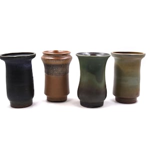 May include: Four handcrafted ceramic vases in varying earth tones. The vases have a cylindrical shape with a flared top. Colors include black, brown, green, and tan. Each vase has a unique texture and finish, showcasing the artistry of the pottery.