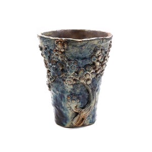 May include: A handcrafted ceramic vase with a textured, dark blue glaze. The vase features a raised floral design with cream-colored blossoms and a brown stem. The vase is approximately 8 inches tall.