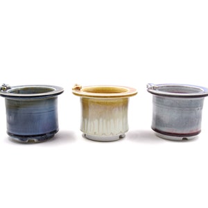 Pode incluir: Três vasos de cerâmica com um design único. Os vasos são de cores diferentes, um é azul, outro amarelo e outro cinza. Cada vaso tem uma pequena abertura no topo para drenagem.