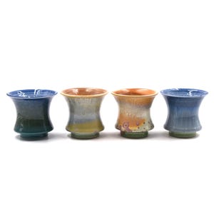 May include: Four ceramic cups with flared rims and tapered bases. The first and fourth cups feature a deep blue to teal gradient glaze. The second cup has a muted green and grey finish, while the third cup displays a warm orange and tan mottled glaze.