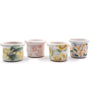 May include: Four small, ceramic flower pots with hand-painted floral designs. The pots feature yellow sunflowers, pink daisies, and yellow and green foliage. Each pot has a different design and color scheme, set against a white background.