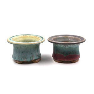 May include: Two small ceramic pots with a flared rim and a textured, cylindrical body. One pot is teal with a yellow rim, the other is brown with a red base. Both have a small, rounded base.
