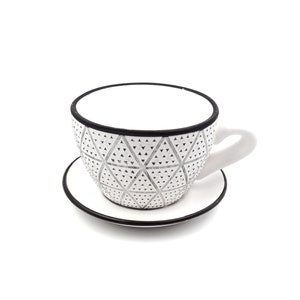 May include: A white ceramic teacup and saucer with a black rim and a geometric pattern of black and silver triangles.