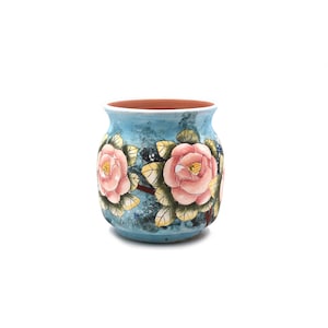 May include: A ceramic vase with a rounded body and a wide opening, featuring a hand-painted design of large pink camellia flowers with green leaves against a mottled light blue background. The interior of the vase shows a natural terracotta color.