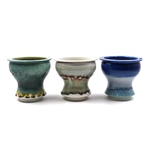 May include: Three ceramic cups with flared rims and tapered bases. The left cup features a mottled teal and green glaze. The center cup has a white, grey, and brown drip glaze. The right cup displays a gradient of dark blue to light blue.